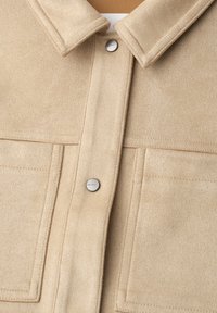 Veste en laine beige avec col pointu, deux poches plaquées devant, et boutons-pression argentés au centre.