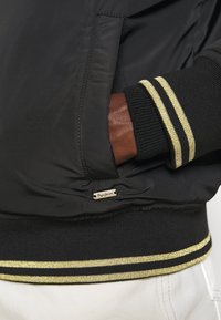 Main dans la poche d'une veste à poignets rayés noir et or, veste noire avec petite étiquette logo métallique Pepe Jeans et pantalon blanc visible.