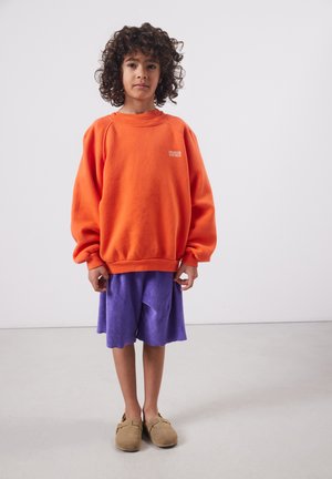 Enfant aux cheveux bouclés debout, portant un sweat-shirt orange surdimensionné, un short violet et des chaussures légères marron clair sur un fond uni.
