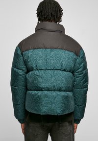 Teal og svart pufferjakke med dekorativt mønster, høy krage og elastiske mansjetter. Har en quiltet tekstur og boksform.
