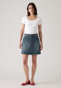 Top blanco de manga corta y escote redondo combinado con una falda de mezclilla azul en forma de A que presenta un cierre de botones en el frente y un dobladillo sin terminar. Zapatos de charol rojos.