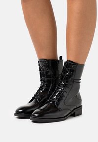 Botas de tobillo de charol negro con un diseño de cordones, que presentan punteras redondas, un acabado texturizado y pequeños tacones de bloque.