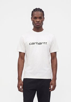 Άνδρας με πλεξούδες, φοράει λευκό t-shirt Carhartt και μαύρο παντελόνι, στέκεται μπροστά σε απλό λευκό φόντο.