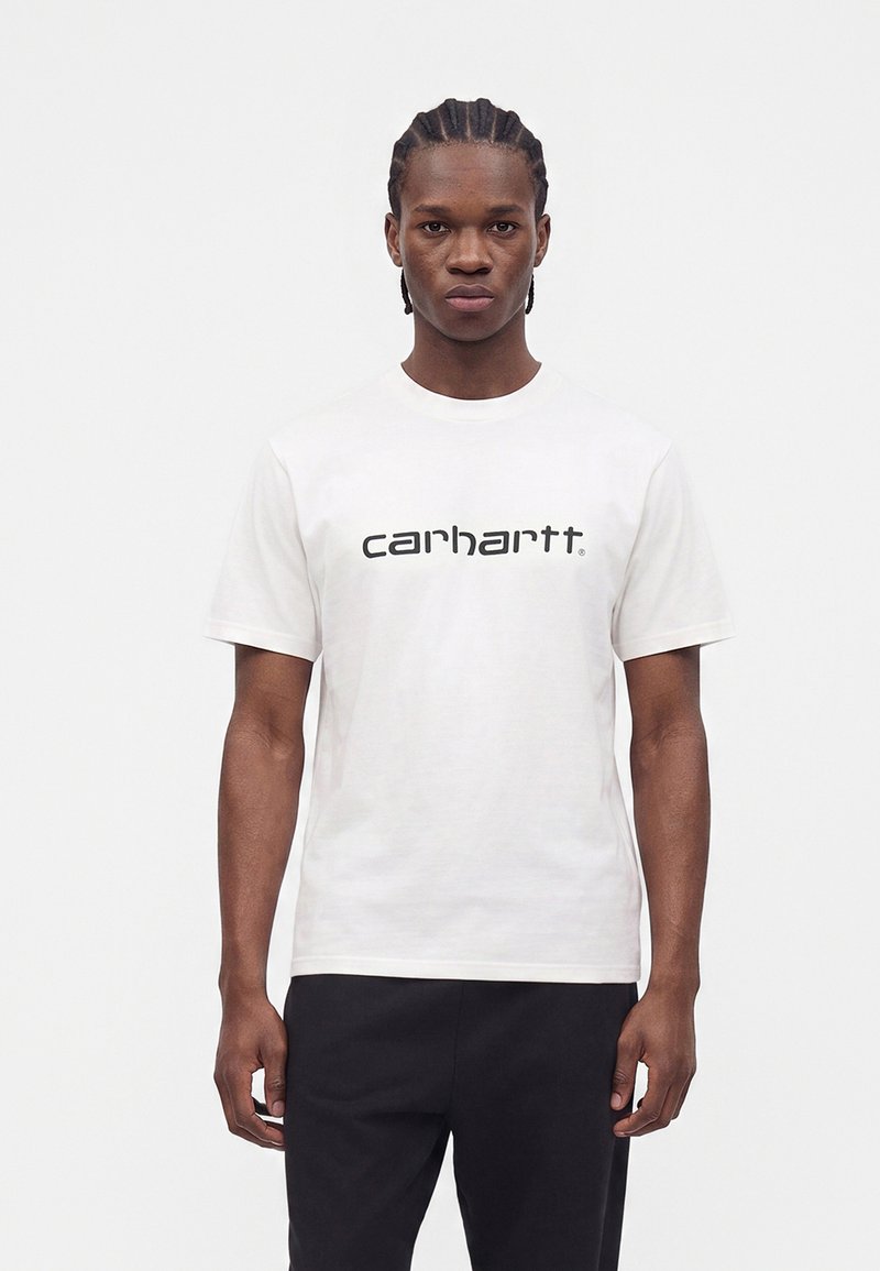 Άνδρας με πλεξούδες, φοράει λευκό t-shirt Carhartt και μαύρο παντελόνι, στέκεται μπροστά σε απλό λευκό φόντο.