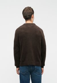 Καφέ πλεκτό πουλόβερ με στρογγυλή λαιμόκοψη, ριμπ μανσέτα και χαλαρή εφαρμογή, συνδυασμένο με μπλε τζιν παντελόνι. Ο υφασμάτινος ιστός φαίνεται απαλός.