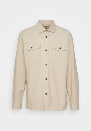 Beige overhemd met knopen, voorzien van twee borstzakken, zwarte knopen en een gestructureerde kraag. De stof lijkt textuur te hebben en het shirt heeft lange mouwen.