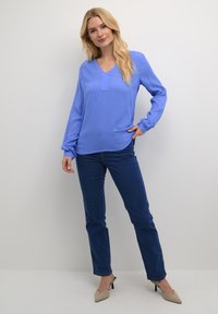 Blusa a maniche lunghe azzurra con scollatura a V e leggere pieghe, abbinata a jeans dritti blu scuro e décolleté beige a punta.