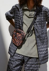 Černá lehká bunda a kalhoty s bílým vzorem loga; olivově zelený polo. Hnědá taška přes tělo s leopardím potiskem, se zipy a popruhem.