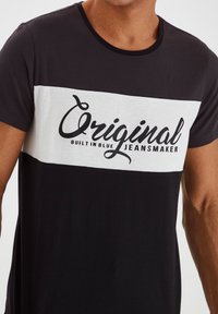 Juoda ir balta trumpa rankovė T-marškiniai su apvaliu iškirpimu ir priekinio vaizdo tekstu "Original Built-in Blue Jeansmaker." Minkšta medvilnės mišinys.