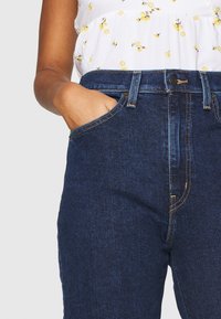 Tmavě modré džíny s vysokým pasem, které mají žluté prošívání a klasický design s pěti kapsami. Kromě toho je ruka opřená v jedné z kapes.