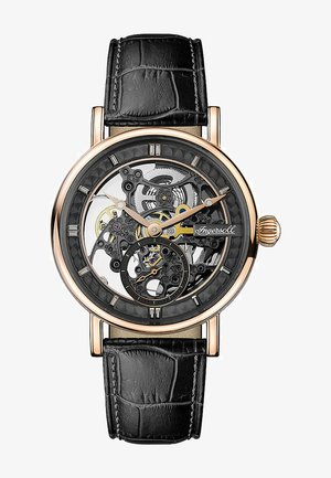 Mechaniczny zegarek z przezroczystym szkieletem na tarczy. Koperta w odcieniu różowego złota, czarny skórzany pasek z tłoczeniem w strukturę krokodyla. Zauważalne, misternie wykonane tryby.