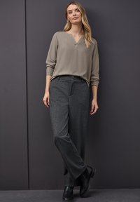 Pitkähihaisessa, V-päisessa taupe-paidassa on yhdistettyt tummanharmaat leveät housut. Housuissa on nappikiinnitys ja sileä pinta. Mustat nilkkurit.