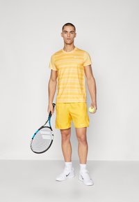 Keltainen raidallinen t-paita ja siihen sopivat shortsit, molemmissa Nike-logo. Valkoiset lenkkarit ja tennismaila käsissä, tennispallo kädessä.