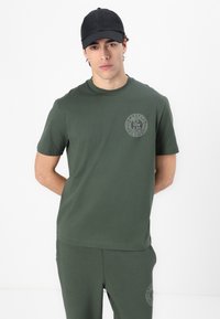 Camiseta de algodón verde oliva con mangas cortas, escote redondo y logo en relieve en el pecho. Combinada con pantalones verdes oliva a juego. Gorro negro.