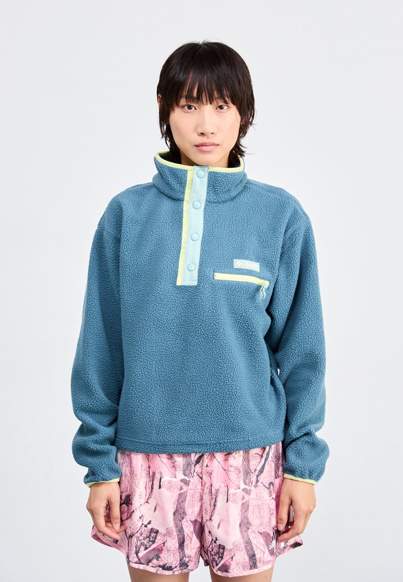 Pulôver de fleece azul com colarinho alto, detalhes em verde claro, botões de pressão e bolso no peito; usado com calções cor-de-rosa com padrão.