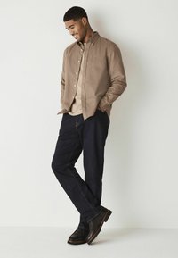 Lichtbruine button-up shirt met een ontspannen pasvorm, gecombineerd met donkerblauwe jeans en zwarte leren schoenen. Strak ontwerp met minimale details.