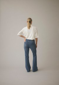 Žena plave punđe u bijelom topu i plavim širokim trapericama stoji s jednom rukom na bokovima, okrenuta prema jednostavnoj svijetloj pozadini.