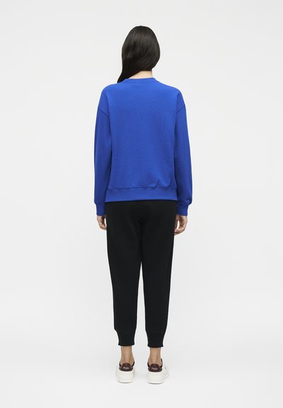 Άτομο με μακριά σκούρα μαλλιά που φορά μπλε φούτερ, μαύρο παντελόνι και λευκά αθλητικά, στέκεται με την πλάτη γυρισμένη σε λευκό φόντο.