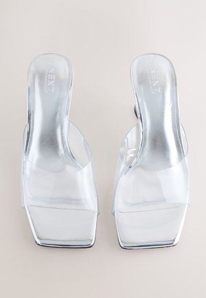 Gennemsigtige højhælede mules med et øvre lag af transparent PVC og et firkantet tå-design. Reflekterende sølvfarvede indlægssåler og stabelhæle fuldender udseendet.