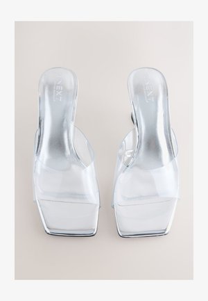 Gennemsigtige højhælede mules med et øvre lag af transparent PVC og et firkantet tå-design. Reflekterende sølvfarvede indlægssåler og stabelhæle fuldender udseendet.