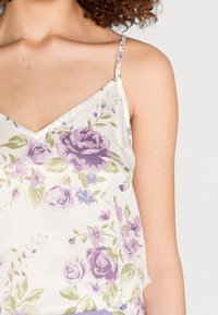 Femme portant un camisole en satin blanc avec un imprimé floral violet et vert, et des fines bretelles sur un fond uni.