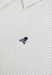 Zoom su un tessuto a strisce beige e bianche con un piccolo teschio e ossa incrociate ricamato in blu navy vicino a un bottone bianco.