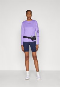 Μπλούζα μακρυμάνικη σε μωβ χρώμα με λογότυπο και σχέδιο στο μανίκι, συνδυασμένη με σκούρα μπλε σορτς ποδηλασίας. Μαύρη ζώνη με θήκη για παγούρι νερού. Λευκά αθλητικά παπούτσια.