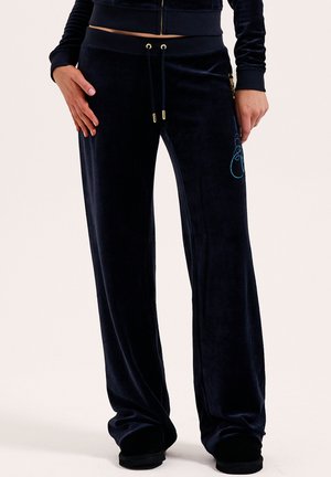 Frau trägt schwarze Samt-Hosen mit weitem Bein, goldenen Zugband und besticktem Design an der Seite und steht mit den Händen an den Seiten.