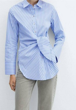 Niebiesko-biała koszula w pionowe paski na guziki z detalem skręconego węzła w talii, noszona z jasnoszarymi spodniami.