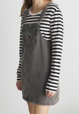 Osoba ubrana w szarą dżinsową sukienkę na szelkach na czarno-białej, paskowanej bluzce z długim rękawem na jednolitym jasnym tle.