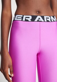 Svetlo roza legice z gladko teksturo, ki imajo črn pas z belim logotipom "UNDER ARMOUR". Prikaz v bližnjem posnetku poudarja podrobnosti prileganja.