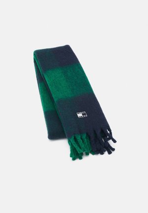 Écharpe en laine présentant un motif à carreaux bleu marine et vert, avec des bords frangés et une petite étiquette logo. Texture douce, forme rectangulaire.