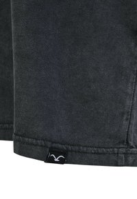 Dunkelgrauer Baumwollstoff mit einer weichen Textur. Verfügt über einen Saum mit einem schwarzen Etikett, das ein weißes Logo zeigt. Keine sichtbaren Muster oder Verzierungen.