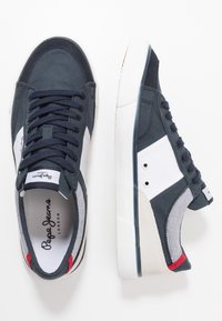 Námořnické plátěné tenisky s bílými a šedými akcenty, texturovaná gumová podrážka, kulatá špička a černé tkaničky. Obsahují logo na jazyku.