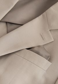 Close-up of beige suit jacket lapel and pocket with visible buttonhole and smooth fabric texture.