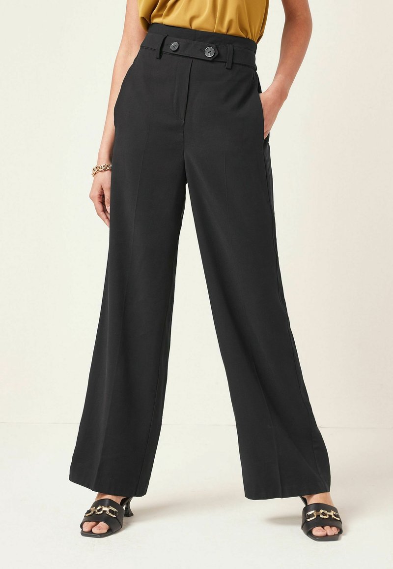 Femme portant un pantalon noir taille haute à jambes larges avec une ceinture boutonnée, associé à des sandales noires à talons ouvertes sur les orteils et un haut couleur moutarde.