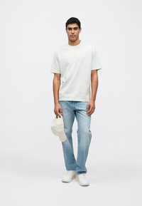 Ανοιχτό γκρι βαμβακερό μπλουζάκι με κοντά μανίκια, συνδυασμένο με ανοιχτό μπλε τζιν. Κρατάει μπεζ καπέλο, φοράει λευκά αθλητικά παπούτσια. Απλός σχεδιασμός.