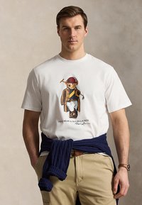 Mann trägt weißes T-Shirt mit Polo-Bären-Motiv, beigen Hosen und dunkelblauen Pullover um die Taille gebunden, steht vor neutralem Hintergrund.