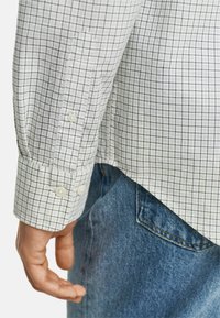 Hvid skjorte med sort rutenet, der har dobbelt knapmanchetter og en buet søm. Kombineret med blå denim jeans.