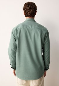 Camicia a maniche lunghe verde con finitura testurizzata, colletto con bottoni e polsini con bottoni, indossata con pantaloni color crema.
