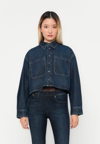 Afgeknotte donkerblauwe denimjack met twee borstzakken, knoopsluiting en gespreide kraag; gedragen over donkere jeans met een gladde textuur.