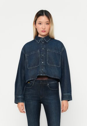 Afgeknotte donkerblauwe denimjack met twee borstzakken, knoopsluiting en gespreide kraag; gedragen over donkere jeans met een gladde textuur.