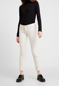 Kvinna som bär en svart långärmad topp, off-white rullade slimbyxor och svarta snörskor, stående mot en enfärgad vit bakgrund.