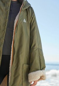 Olivengrøn parka med blødt beige fleecefor, lynlås lukning og orange detaljer; har et trekantet logo og sidelommer.