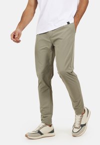 Jasnooliwkowe, zwężane spodnie wykonane z gładkiego materiału, w połączeniu z białymi i szarymi butami sportowymi z teksturowaną podeszwą i akcentującymi kolorami.