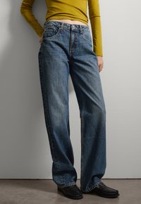 Jeans de mezclilla de lavado claro con un corte de pierna ancha, que cuentan con bolsillos delanteros y cierre de botón. Combinados con una blusa de manga larga mostaza y zapatos negros.
