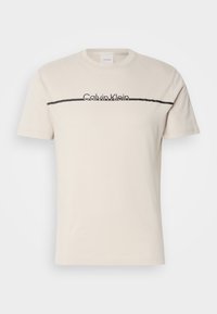 T-shirt beige clair à manches courtes avec col rond, comportant une fine ligne horizontale noire et le texte « Calvin Klein » sur la poitrine.