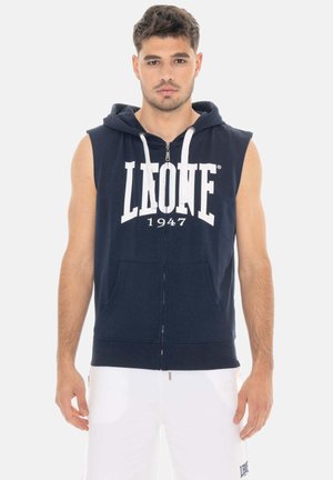 Un homme porte un sweat à capuche bleu marine sans manches, zippé, avec des cordons blancs et un lettrage bien visible, assorti à un short blanc, sur un fond uni.