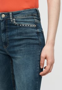 Κοντινό από τη μέση, που φοράει σκούρα μπλε τζιν με στρας στην άκρη της τσέπης και πορτοκαλί τοπ, με το χέρι ακουμπισμένο στο πλάι.