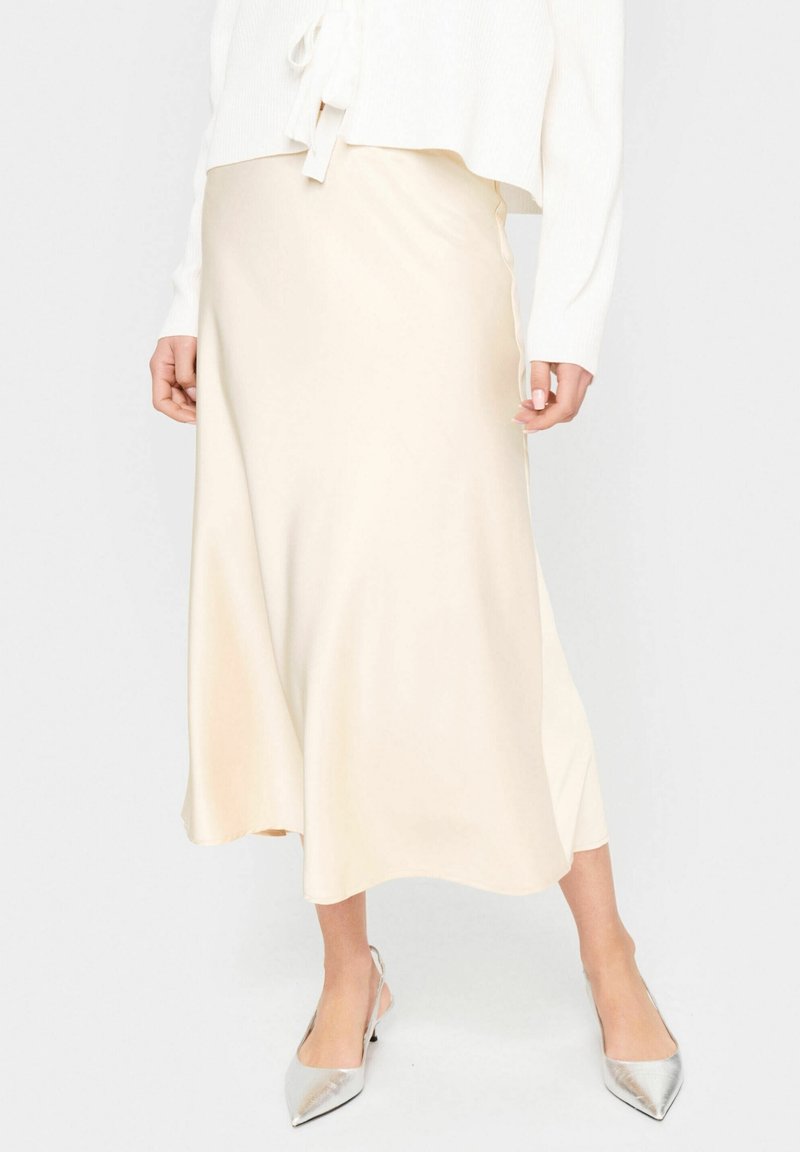 Jupe midi en satin beige portée avec un haut blanc à manches longues et des talons slingback argentés à bout pointu, visible de la mi-torse aux pieds.